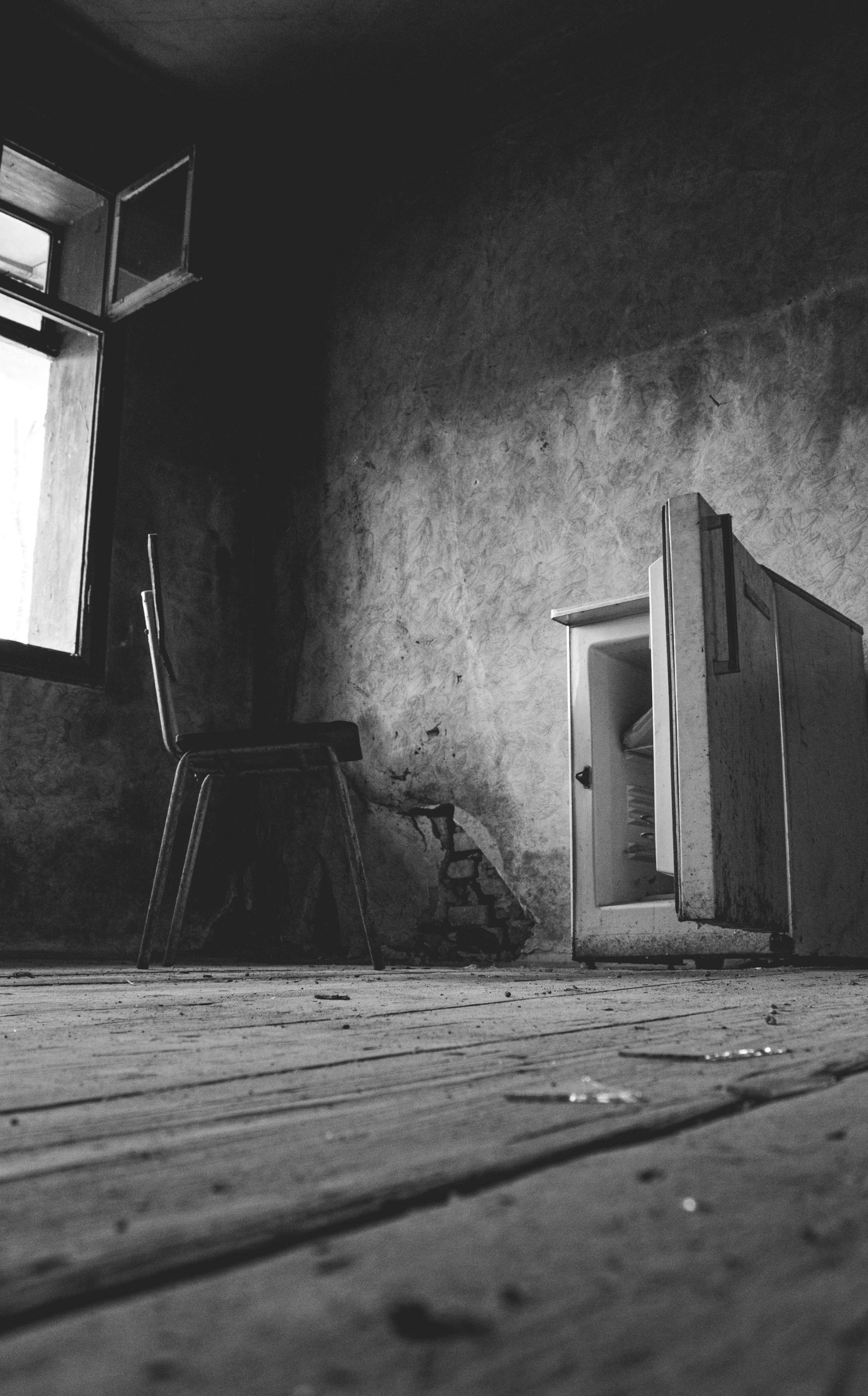 A dramatic black and white photo of an abandoned room featuring a chair and open freezer. Une photo dramatique en noir et blanc d'une pièce abandonnée avec une chaise et un congélateur ouvert.