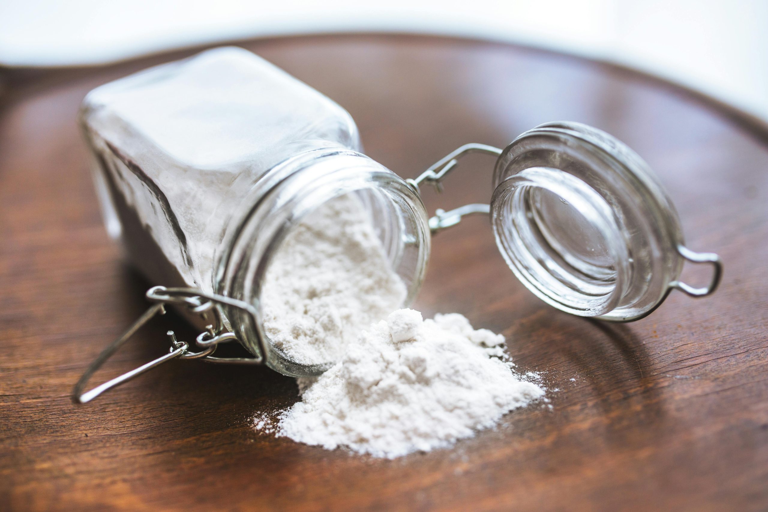 Glass jar tipped over with white flour spilling onto a wooden surface, ideal for baking themes. Un bocal en verre renversé avec de la farine blanche se déversant sur une surface en bois, idéal pour les thèmes de pâtisserie.